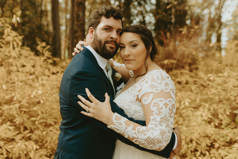 bride in lace dress with groom in charcoal suit with arms around each other and fall greenery behind them