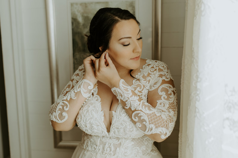 bride in lace dress with deep v-neck putting in earrings by farmhouse window